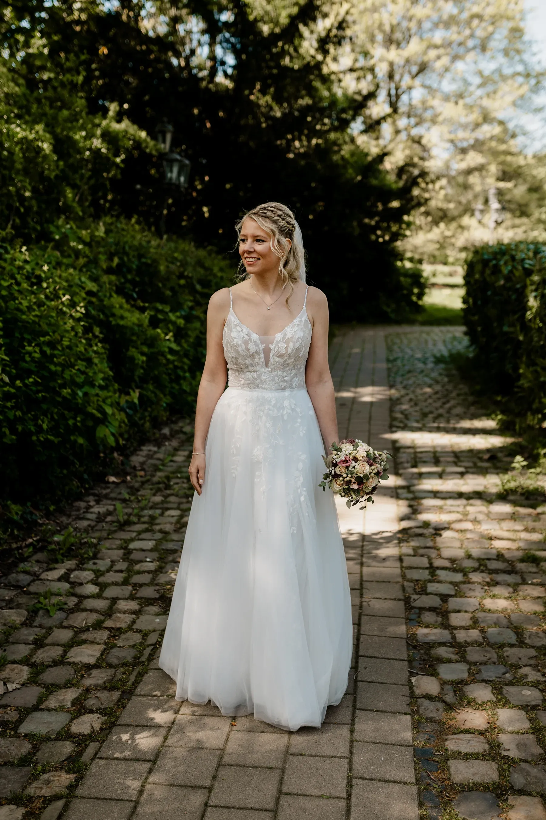 Braut in einem Spitzenkleid mit Blumenstrauß auf einem Gartenweg